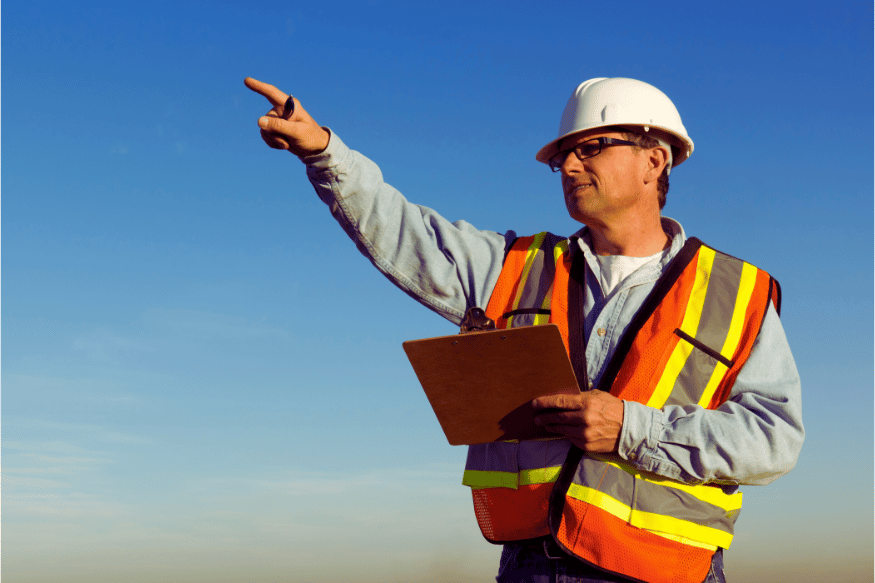 Man wearing high-visibility clothing and a hard hat pointing while holding a clipboard on a worksite.